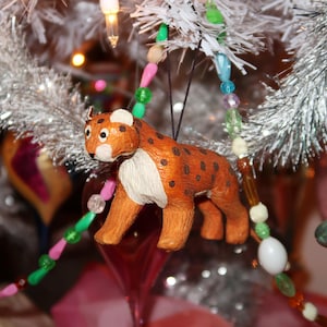 May include: A handcrafted Christmas tree ornament shaped like a leopard. The ornament is orange with brown spots and white accents. It hangs from a beaded garland with green, pink, and yellow beads. The background includes a white Christmas tree and other ornaments.