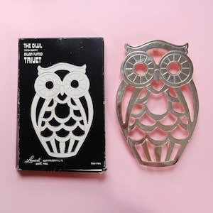 May include: A silver-plated owl trivet and its original box. The trivet is in the shape of an owl with cut-out details. The box is black with white text that reads "THE OWL SILVER PLATED TRIVET".