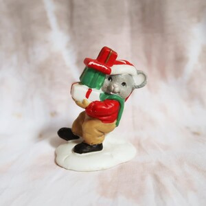 May include: A ceramic Christmas mouse figurine carrying wrapped gifts. The mouse wears a red jacket, green scarf, and a Santa hat. The gifts are green, white, and red. The figurine is standing on a white base.