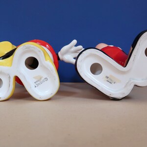 May include: Two ceramic figurines of Mickey Mouse, one in yellow and one in red, with the Disney logo on the bottom.