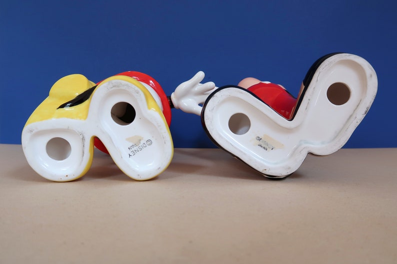 May include: Two ceramic figurines of Mickey Mouse, one in yellow and one in red, with the Disney logo on the bottom.