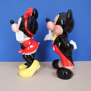 May include: A pair of ceramic figurines of Mickey Mouse and Minnie Mouse. Mickey is wearing a black tuxedo with a red bow tie and red trousers. Minnie is wearing a red dress with white polka dots and a yellow bow. The figurines are standing side by side on a light brown surface.