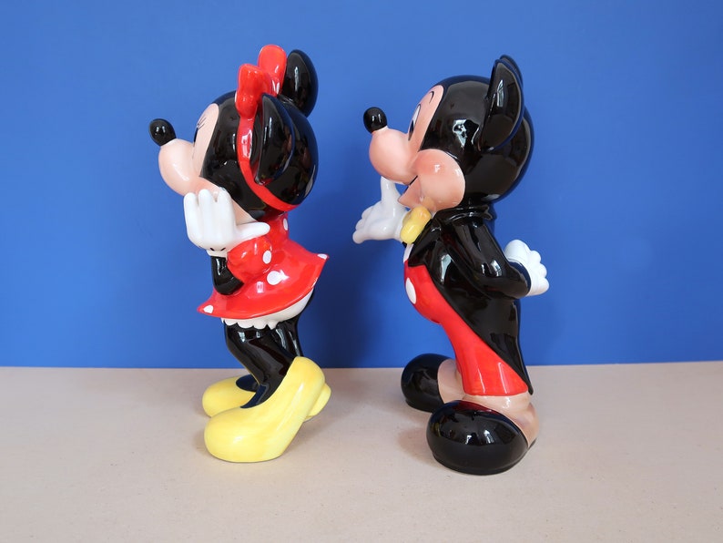 May include: A pair of ceramic figurines of Mickey Mouse and Minnie Mouse. Mickey is wearing a black tuxedo with a red bow tie and red trousers. Minnie is wearing a red dress with white polka dots and a yellow bow. The figurines are standing side by side on a light brown surface.