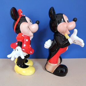 May include: A ceramic figurine set of Mickey Mouse and Minnie Mouse. Mickey is wearing a black tuxedo with a red bow tie and red trousers. Minnie is wearing a red dress with white polka dots and a yellow bow.