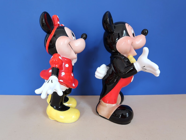 May include: A ceramic figurine set of Mickey Mouse and Minnie Mouse. Mickey is wearing a black tuxedo with a red bow tie and red trousers. Minnie is wearing a red dress with white polka dots and a yellow bow.