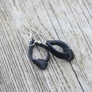 Puede incluir: Un par de pendientes de cuero negro con ganchos de plata. Los pendientes tienen forma de bucle con un nudo en la parte inferior.