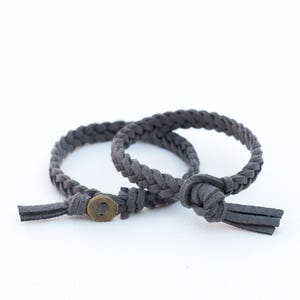 May include: Two gray braided leather bracelets with a metal button closure and tassels.