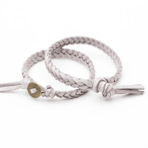 Puede incluir: Dos pulseras de cuero trenzado con un pequeño botón de latón. Las pulseras son de color gris claro.