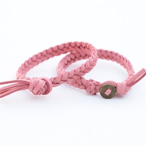 May include: Two pink braided leather bracelets. One bracelet has a tassel, and the other has a bronze-colored button clasp. The bracelets are on a white background. These accessories are perfect for adding a touch of color to any outfit.