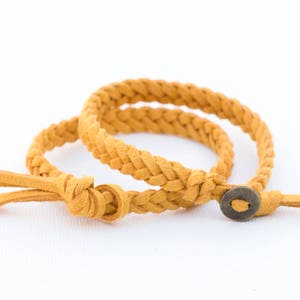 Può includere: Due braccialetti in pelle intrecciata di colore giallo senape. Un braccialetto ha una chiusura a bottone rotonda piccola in ottone.