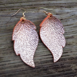 May include: A pair of rose gold leaf-shaped earrings with a textured finish. The earrings are made of leather and have gold-toned hooks.