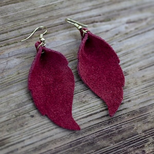 Puede incluir: Un par de pendientes de hoja de gamuza color burdeos con ganchos dorados.