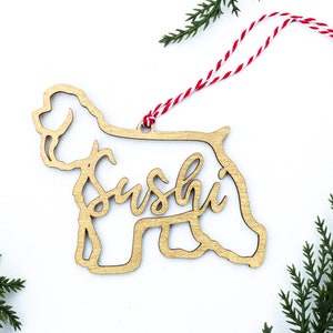 May include: A wooden Christmas ornament shaped like a dog with the name "Sushi" written inside the silhouette. The ornament is gold and has a red and white string attached.
