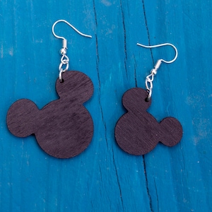 May include: A pair of black wooden earrings shaped like Mickey Mouse heads. The earrings are hanging from silver hooks on a blue wooden background.