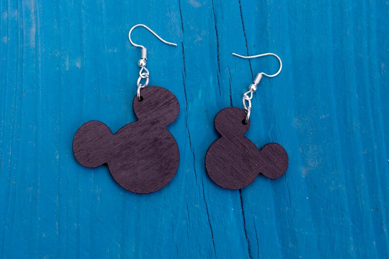 May include: A pair of black wooden earrings shaped like Mickey Mouse heads. The earrings are hanging from silver hooks on a blue wooden background.