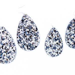 Puede incluir: Un conjunto de cuatro pendientes de gota de purpurina plateada. Los pendientes son de diferentes tamaños y todos están hechos con un material de purpurina plateada.