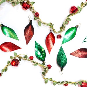 Könnte beinhalten: Eine Sammlung von grünen und roten Lederohrringen in Blattform. Die Ohrringe sind mit Glitzer verziert und haben eine glänzende Oberfläche. Die Ohrringe werden auf einem weißen Hintergrund mit einer Girlande aus grünen Blättern und roten Glöckchen präsentiert.