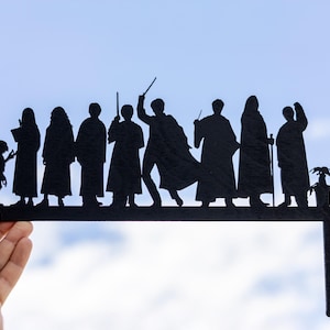 May include: Black silhouette paper cutouts of characters from the Harry Potter series, including Harry Potter, Ron Weasley, Hermione Granger, and others. The cutouts are arranged in a line on a black base with a small plant in a pot on the right side.