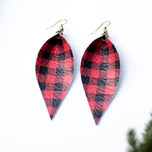 May include: Two leaf-shaped earrings featuring a red and black buffalo check pattern. They have gold-coloured ear wires and are displayed against a white backdrop. These earrings are a festive accessory.