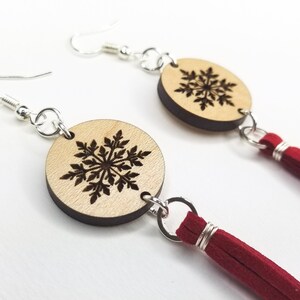 May include: A pair of wooden snowflake earrings with red tassel accents. The earrings are round and feature a laser-cut snowflake design.