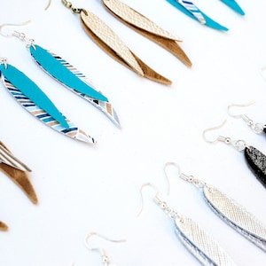 May include: Assortment of handmade leather earrings in various colours and patterns. The earrings feature a long, teardrop shape with a layered design. Some earrings have a silver glitter finish, while others have a striped or geometric pattern.