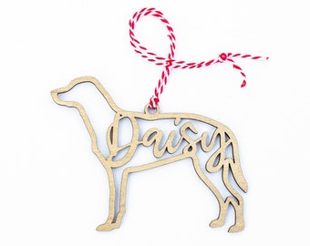 May include: Wooden dog ornament with the name "Daisy" cut out in the shape of a dog. The ornament is attached to a red and white string.