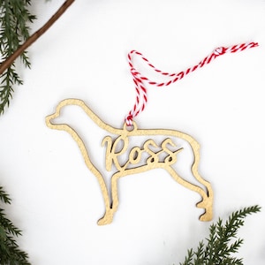 May include: A wooden dog ornament with the name "Ross" cut out in the middle. The ornament is a light brown color and is hanging from a red and white string.