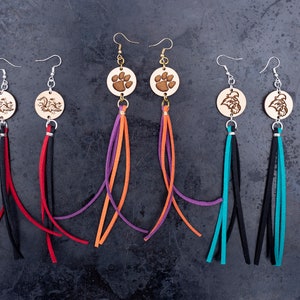 May include: Six pairs of dangle earrings with wooden charms and colourful tassels. The charms feature a paw print, a dragon, and a bird. The tassels are in shades of purple, orange, turquoise, black, and red.