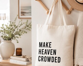 May include: A cream-colored canvas tote bag featuring the black printed text "MAKE HEAVEN CROWDED" in a bold, sans-serif font. The bag has two matching fabric shoulder straps and is hanging from a wooden wall-mounted rack.