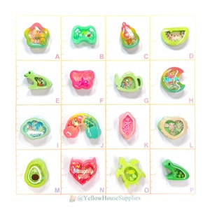 May include: Assortment of 16 colorful resin charms with various designs, including a frog, a butterfly, a teapot, a crescent moon, a rainbow, a heart, an avocado, and a turtle. Each charm is filled with glitter and other small decorative elements.