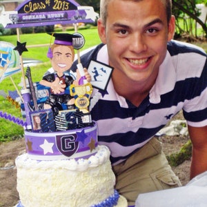 K&ouml;nnte beinhalten: Eine mehrst&ouml;ckige Torte mit wei&szlig;er Glasur und violetten Akzenten, gekr&ouml;nt von einem Class of 2013 Gonzaga Rugby Schild, einer Comic-Absolventenfigur mit Football, einem goldenen MVP-Pokal sowie verschiedenen sternf&ouml;rmigen Ausschnitten und Fotos.