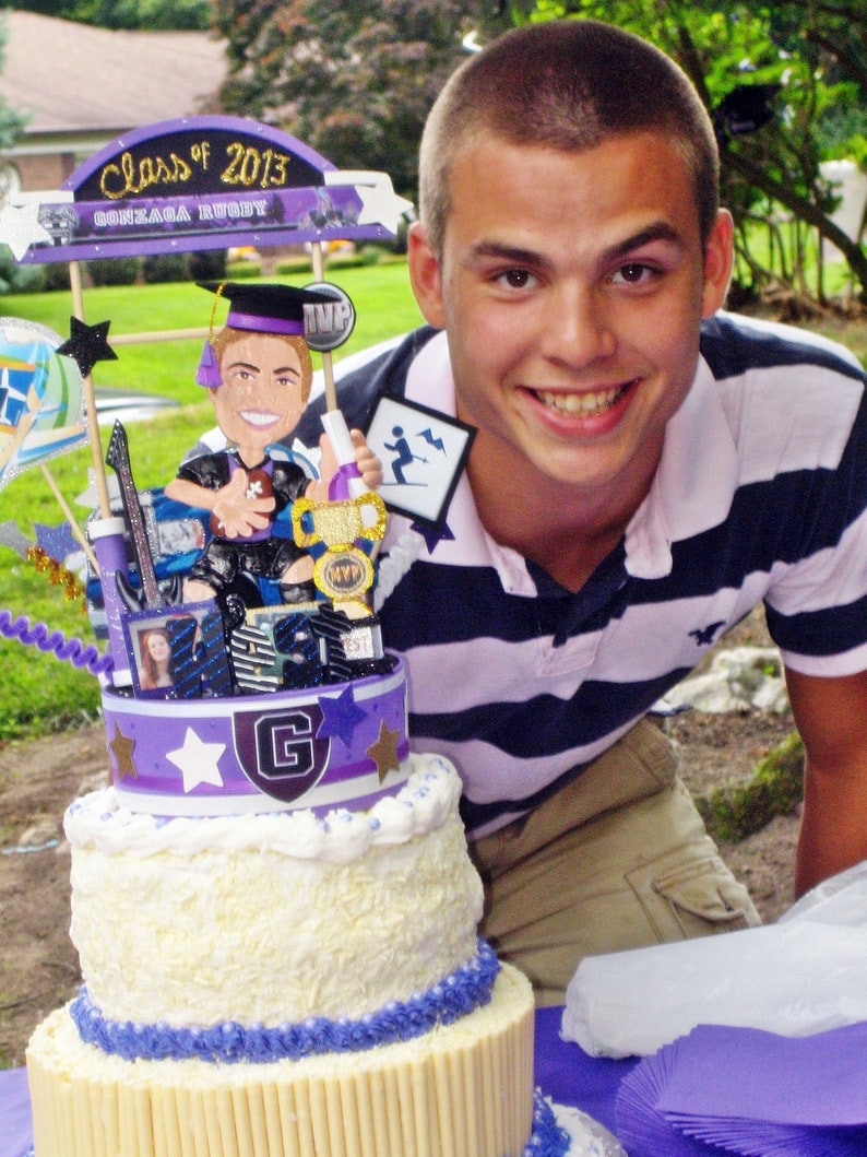 K&ouml;nnte beinhalten: Eine mehrst&ouml;ckige Torte mit wei&szlig;er Glasur und violetten Akzenten, gekr&ouml;nt von einem Class of 2013 Gonzaga Rugby Schild, einer Comic-Absolventenfigur mit Football, einem goldenen MVP-Pokal sowie verschiedenen sternf&ouml;rmigen Ausschnitten und Fotos.