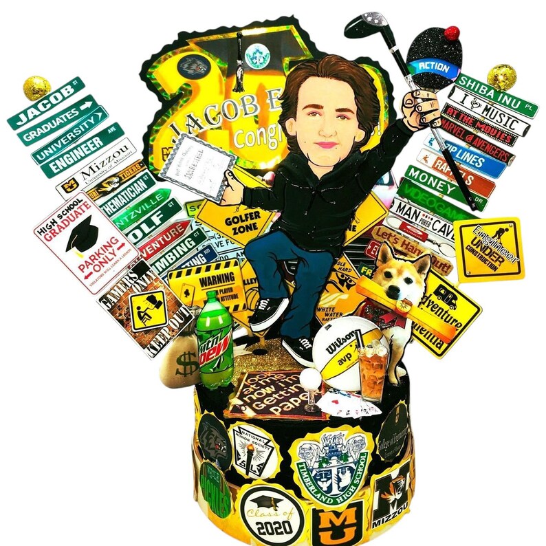 K&ouml;nnte beinhalten: Ein Tortenaufleger mit einer Cartoon-Illustration eines Absolventen in schwarzer Jacke und blauer Jeans, umgeben von Miniaturschildern mit Texten wie Jacob, Mizzou und Engineer, einem Hund, einem Golfschl&auml;ger, einer Limonadenflasche und einem Volleyball.
