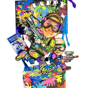 May include: A vibrant paintball-themed cake featuring two cartoon figures, one with a paintball gun. The cake is adorned with paint splatters and the words "Happy 10th Birthday". A sign reads "Reserved Parking for Paintballers".