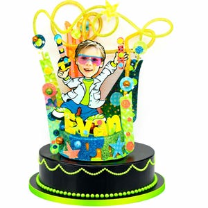 Könnte beinhalten: Ein dekoratives Mittelstück mit einer Cartoon-Illustration eines Kindes mit rosagetönten Brillen, einer weißen Jacke und einer blauen Hose. Der Name Evan ist in gelben Buchstaben dargestellt. Die Basis ist schwarz mit grünem Perlenbesatz.