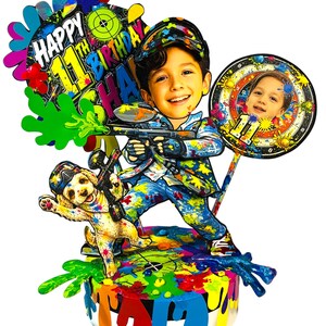 May include: A multi-layered cake topper featuring a cartoon-style child holding a paintball gun, a puppy, and a circular sign with a photo. The text reads Happy 11th Birthday. The design includes vibrant paint splatters in blue, red, green, and yellow.