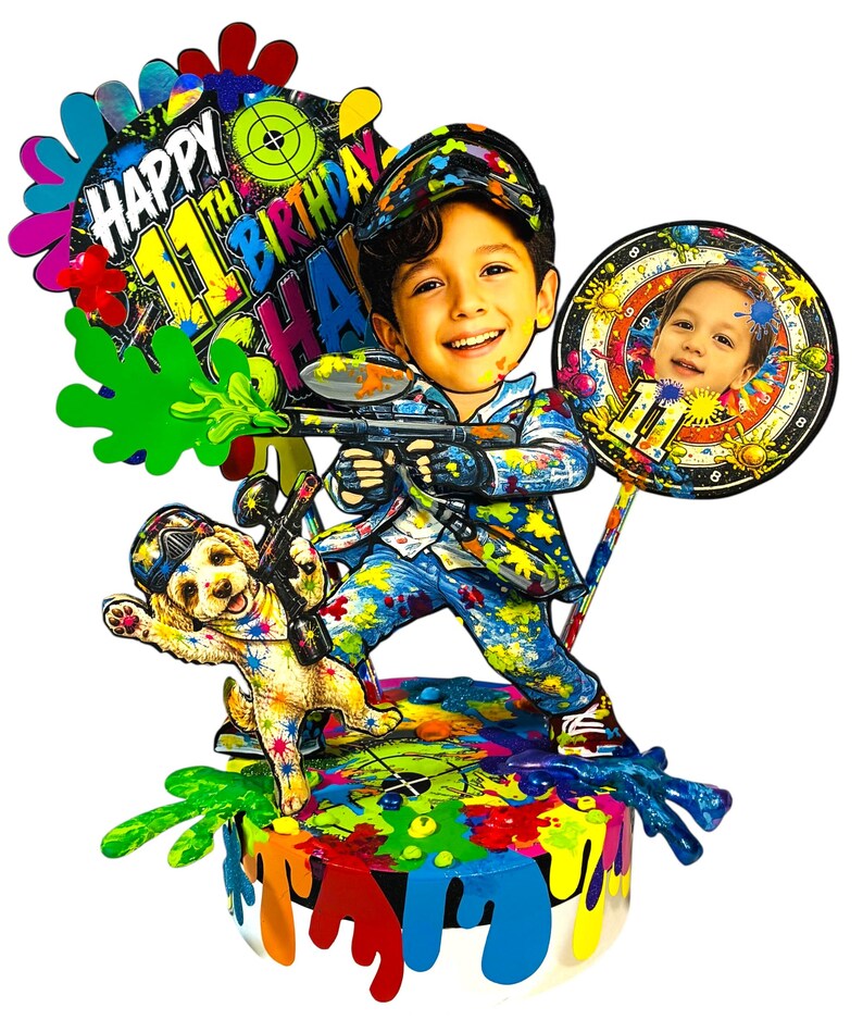 May include: A multi-layered cake topper featuring a cartoon-style child holding a paintball gun, a puppy, and a circular sign with a photo. The text reads Happy 11th Birthday. The design includes vibrant paint splatters in blue, red, green, and yellow.
