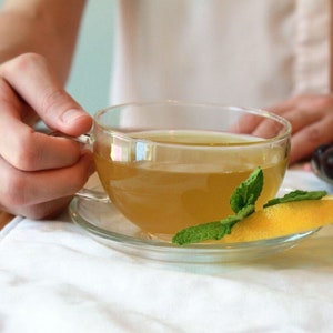 May include: A glass cup of green tea with a lemon wedge and fresh mint leaves on a white saucer.