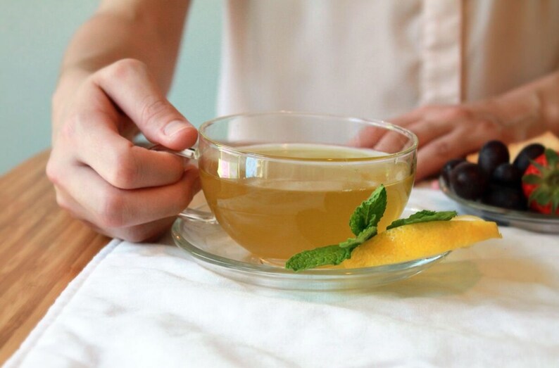 May include: A glass cup of green tea with a lemon wedge and fresh mint leaves on a white saucer.