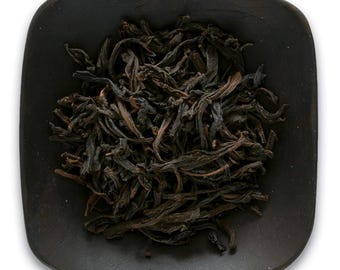 May include: Loose black tea leaves in a small black ceramic bowl. The tea leaves are dark brown and have a slightly curled shape.