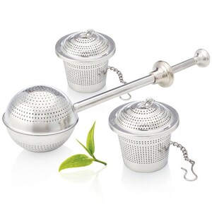 May include: A stainless steel tea infuser set with a sphere and two cup-shaped strainers. The sphere infuser has a long handle with a squeeze mechanism. Each strainer has a chain and a lid. A sprig of green tea leaves is next to the infusers.