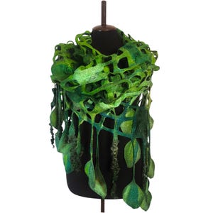 Puede incluir: Bufanda de fieltro verde hecha a mano con un diseño único de tejido abierto. La bufanda presenta diferentes tonos de verde, con detalles en forma de hoja colgando de la parte inferior. La bufanda se exhibe en un maniquí negro, mostrando su intrincada textura y estilo artístico.