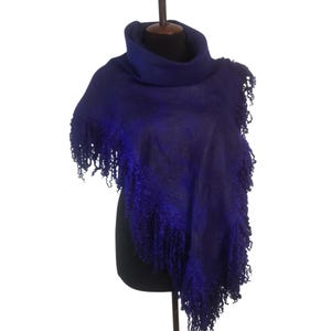 May include: A deep indigo poncho with a cowl neck and fringed edges. The poncho is displayed on a black mannequin, showcasing its asymmetrical design and textured fabric. The fringe adds a decorative element to the garment.