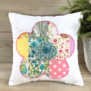 May include: A white pillow with a patchwork flower design. The flower is made of different floral fabrics in shades of pink, yellow, blue, and green. The flower has a large blue button in the center.