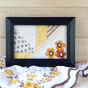May include: A framed patchwork of fabric scraps in shades of yellow, white, and brown with a floral pattern. The fabric is arranged in a geometric pattern and includes lace trim.