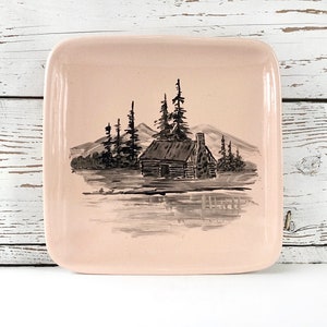 May include: A pink square ceramic dish with a black and white illustration of a cabin in the woods. The cabin is surrounded by trees and mountains.