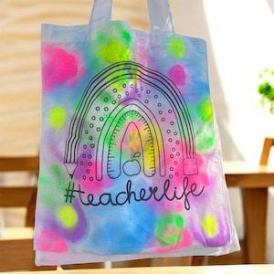 May include: A clear tote bag with a colorful, airbrushed design. The design features a rainbow, pencil, and apple, with the text "#teacherlife" in black script. The background has splashes of pink, blue, green, and yellow.