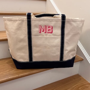 Può includere: Una grande borsa tote in tela bianca con rifiniture nere e un monogramma ricamato rosa "MB". La borsa è appoggiata su una scala di legno.