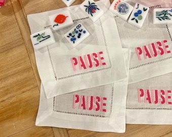 May include: Four white linen napkins with pink embroidered text that reads "PAUSE" on each napkin. The napkins are arranged in a stack with the top napkin folded in half. There are several small, colorful tiles scattered around the napkins.