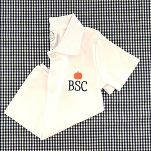 May include: White short-sleeved polo shirt with an embroidered pumpkin and the initials "BSC" in navy blue.